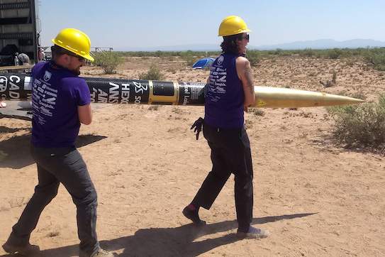 two students wearing hardhats carrying a rocket