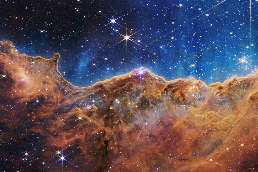 an undulating, translucent star-forming region in the Carina Nebula is shown in this Webb image, hued in ambers and blues; foreground stars with diffraction spikes can be seen, as can a speckling of background points of light through the cloudy nebula