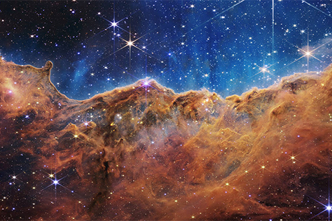 an undulating, translucent star-forming region in the Carina Nebula is shown in this Webb image, hued in ambers and blues; foreground stars with diffraction spikes can be seen, as can a speckling of background points of light through the cloudy nebula