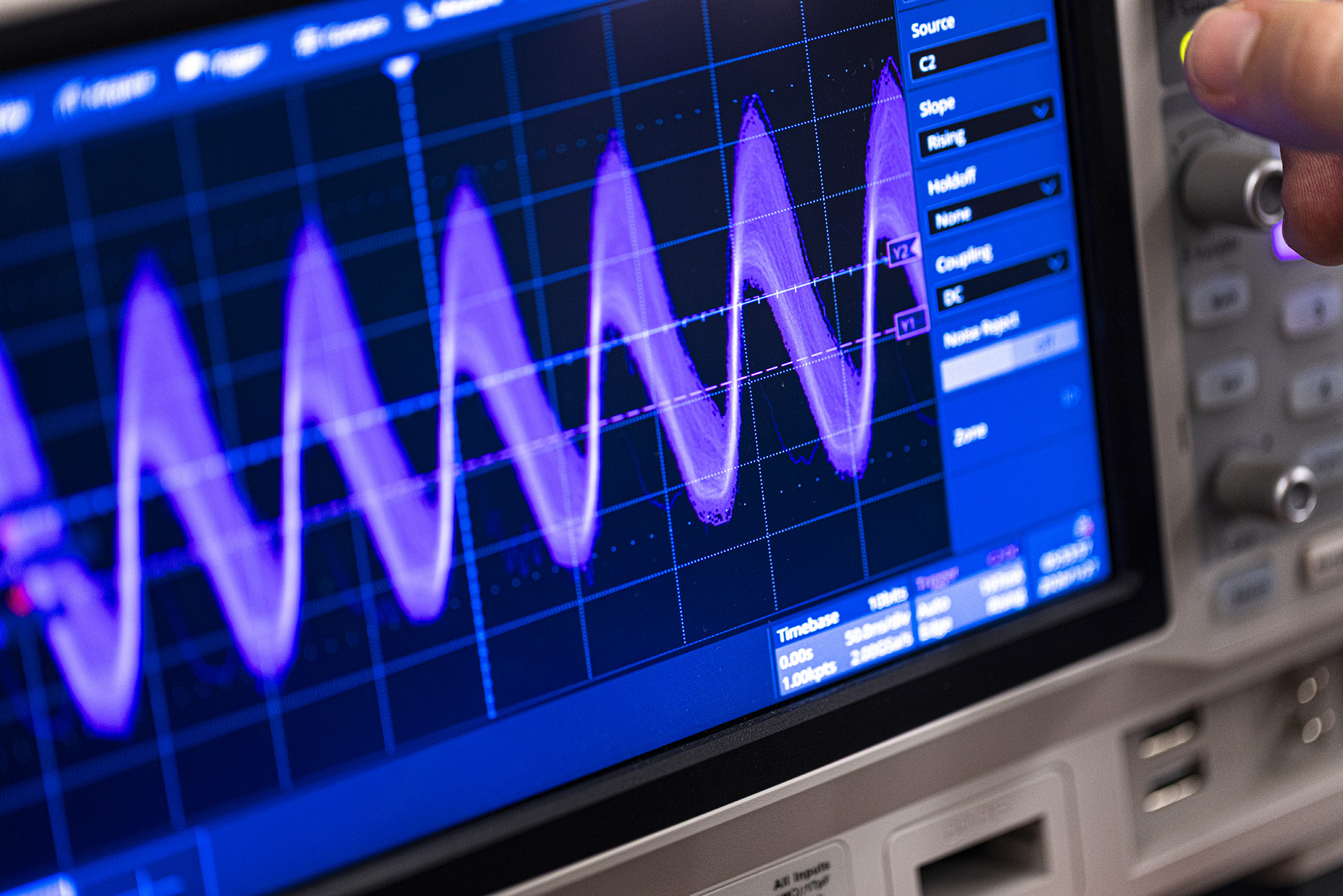 Close-up of an oscilloscope screen displaying a purple waveform with a hand adjusting the controls