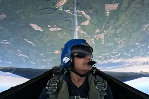 Pilot in a jet cockpit flying upside down over a landscape with forests and fields.