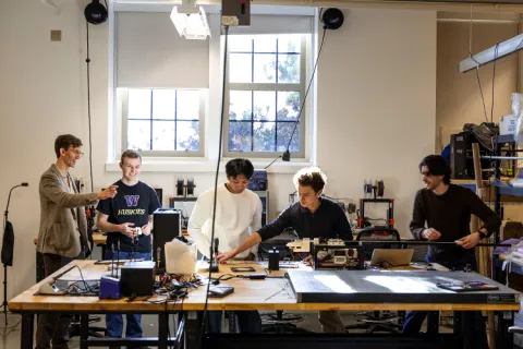 Five people working together at a table in a workshop with equipment and tools