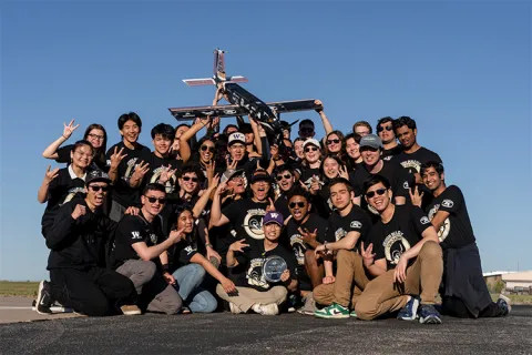 Group of people pose outdoors with a model aircraft under a clear blue sky.